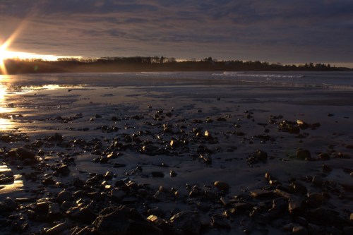 Sunset over a rocky beach, with shimmering water and dark clouds in the sky, creating a serene coastal scene.