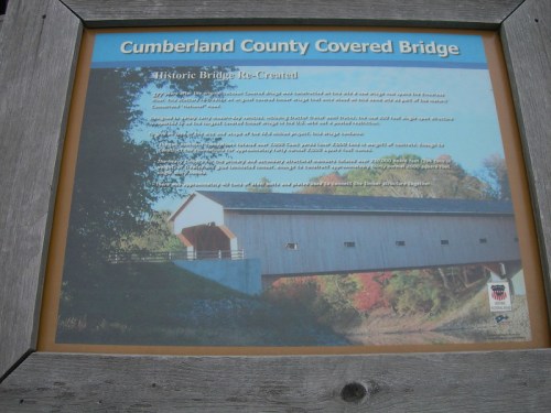 Sign about the Cumberland County Covered Bridge, detailing its history and reconstruction. Scenic bridge in the background.
