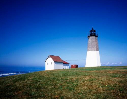 A tall lighthouse beside a small white building, set against a clear blue sky and calm ocean. Green grass in the foreground.