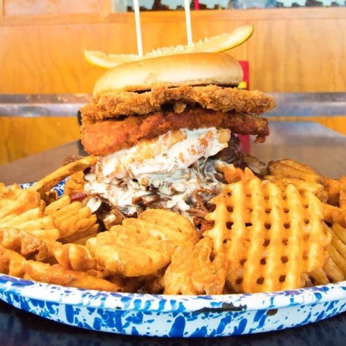 A towering burger with fried chicken, sauce, and a lemon slice, served with a variety of crispy waffle fries.