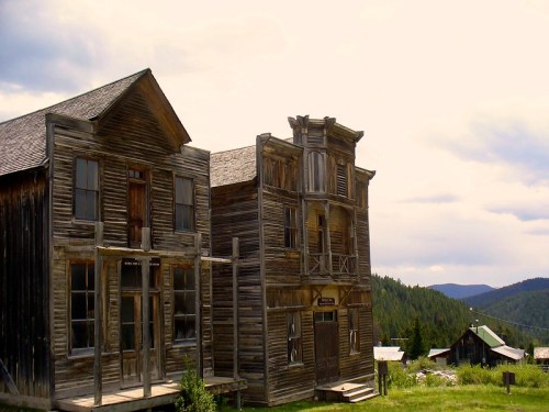 Abandoned wooden buildings in a ghost town, surrounded by trees and mountains under a cloudy sky.