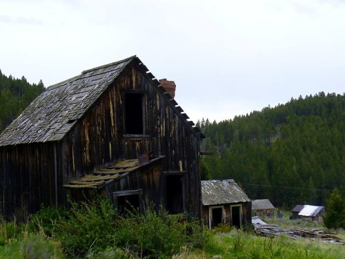 An old, weathered wooden house with broken windows, surrounded by lush green trees and a cloudy sky.