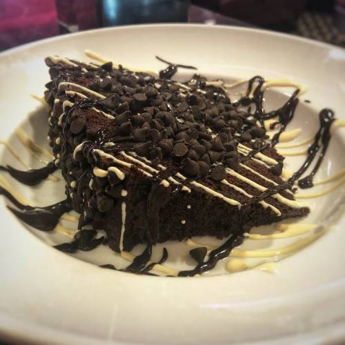 A slice of chocolate cake topped with chocolate chips and drizzled with white and dark chocolate sauces.