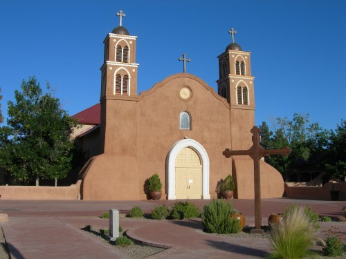 Historic adobe church with two towers, crosses, and a clear blue sky in the background. Desert landscaping surrounds the building.