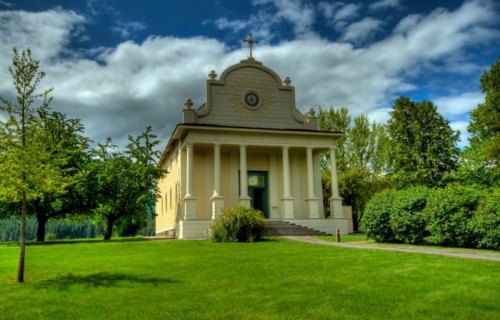 A charming church with a cross, surrounded by lush greenery and trees under a partly cloudy sky.