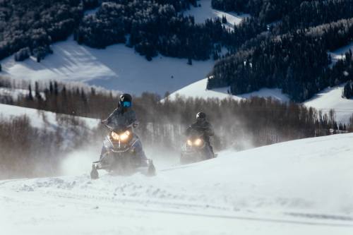 Two snowmobiles ride through a snowy landscape, kicking up powder against a backdrop of mountains and trees.