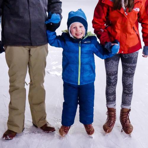 A smiling child in a blue jacket stands on ice, holding hands with two adults in winter clothing.
