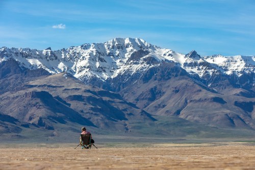 A person sits on a chair, gazing at snow-capped mountains under a clear blue sky.