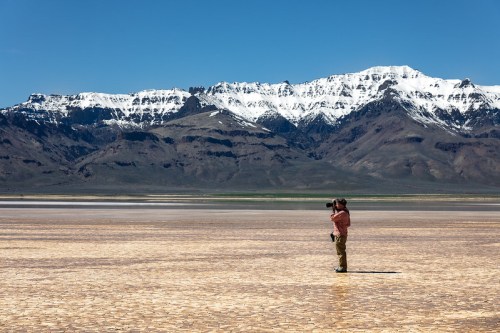 A photographer stands on a dry, cracked salt flat with snow-capped mountains in the background under a clear blue sky.