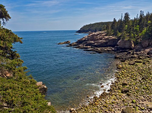 A serene coastal scene with rocky shores, green trees, and calm blue waters under a clear sky.