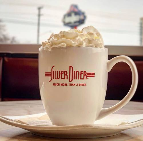 A white mug with "Silver Diner" logo, topped with whipped cream, sits on a table with a blurred diner background.