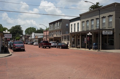 A brick-paved street lined with historic buildings and parked cars under a partly cloudy sky.