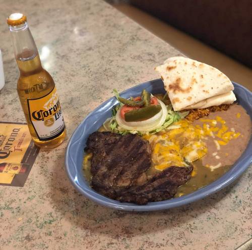 A plate of grilled steak with rice, beans, cheese, tortillas, and a side salad, accompanied by a bottle of Corona beer.
