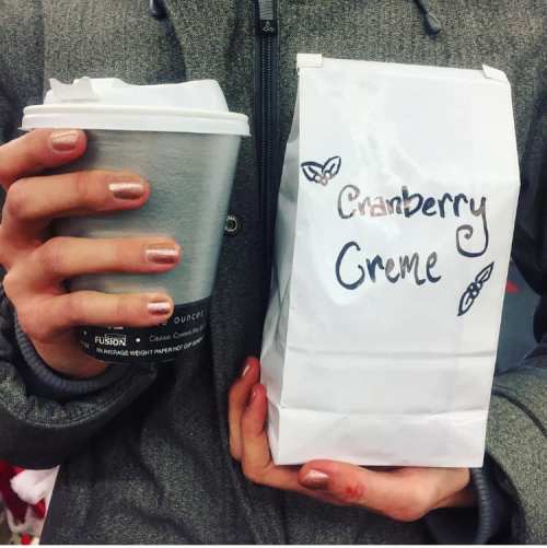 A person holds a coffee cup and a bag labeled "Cranberry Crème," showcasing their stylish nails.