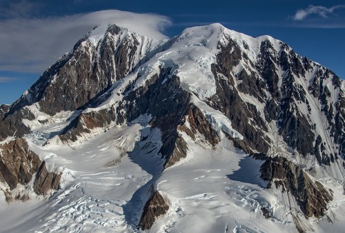 Aerial view of a snow-covered mountain range with rugged peaks and glaciers under a clear blue sky.