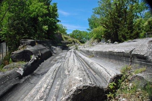 A rocky, sloped landscape with deep grooves, surrounded by greenery and blue sky.
