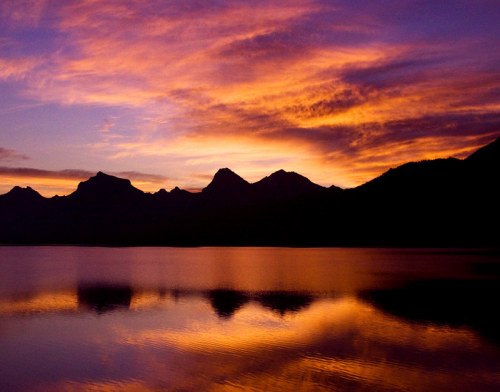 A serene lake at sunset, reflecting vibrant purple and orange hues against silhouetted mountains.