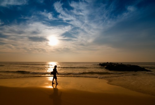 Silhouette of a person walking along a beach at sunset, with waves gently lapping at the shore and clouds in the sky.