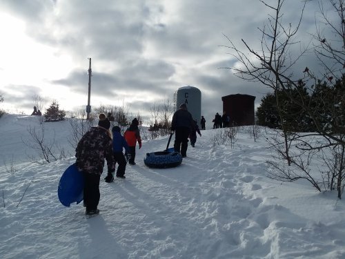 A group of people walking up a snowy hill, carrying sleds, with a cloudy sky and trees in the background.