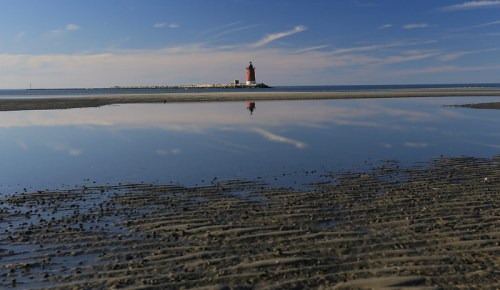 A red lighthouse stands on a rocky shore, reflecting in calm water under a blue sky with wispy clouds.