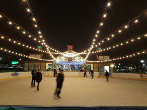 A lively ice skating rink at night, adorned with string lights, with skaters gliding on the ice and a cozy building in the background.