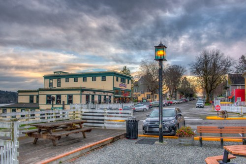 A scenic view of a waterfront area with a restaurant, parked cars, and a lamppost under a cloudy sky.