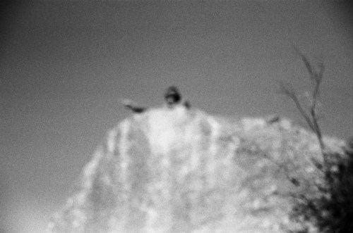 A blurred black-and-white image of a large statue on a rocky outcrop, with a clear sky in the background.
