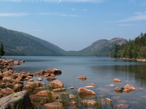 A serene lake surrounded by mountains, with rocky shores and lush greenery under a clear blue sky.