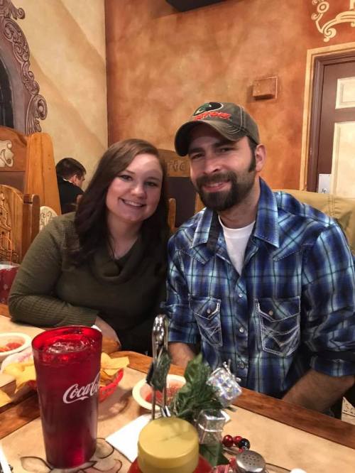 A couple smiles together at a restaurant, with food and drinks on the table in a cozy setting.