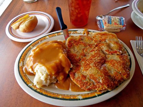 A plate with crispy fried chicken, gravy-covered mashed potatoes, and a side of bread on a table.