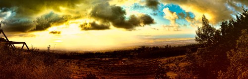 A panoramic view of a landscape at sunset, featuring rolling hills, fields, and dramatic clouds in the sky.