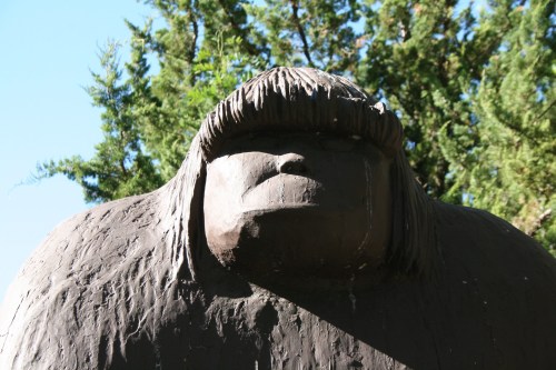 A large, dark sculpture of a figure with a prominent face and long hair, surrounded by trees against a clear sky.