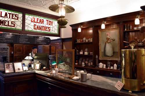 A vintage candy shop interior with wooden counters, glass jars, and a staff member behind the counter.