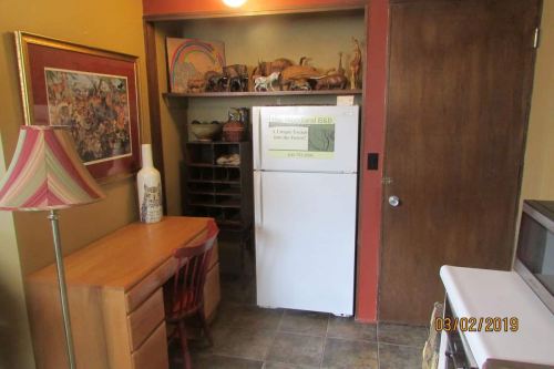 A cozy kitchen nook featuring a desk, chair, refrigerator, and shelves with decorative items and a framed artwork.
