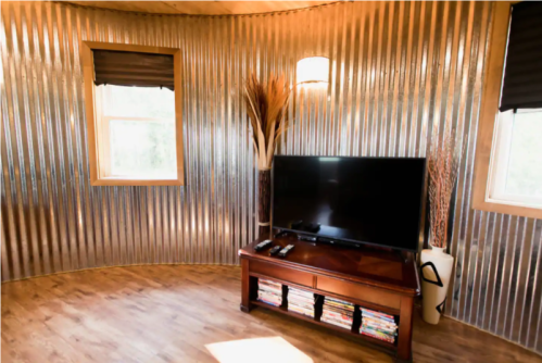 A cozy room with corrugated metal walls, a TV on a wooden stand, and large windows letting in natural light.