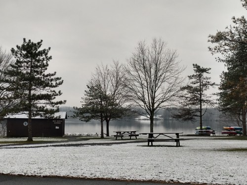 A snowy landscape by a lake, featuring bare trees, picnic tables, and colorful kayaks along the shore.