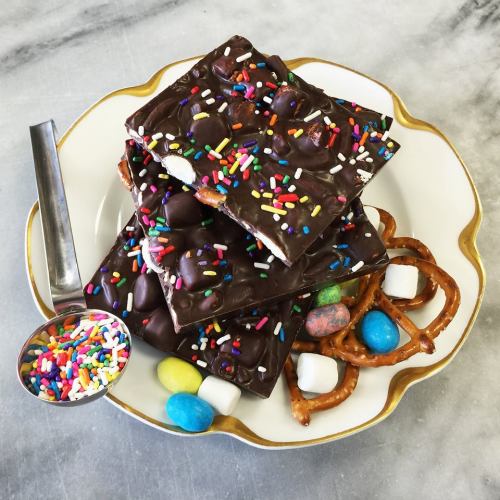 A plate of chocolate bark topped with colorful sprinkles, pretzels, and candy, with a spoonful of sprinkles beside it.