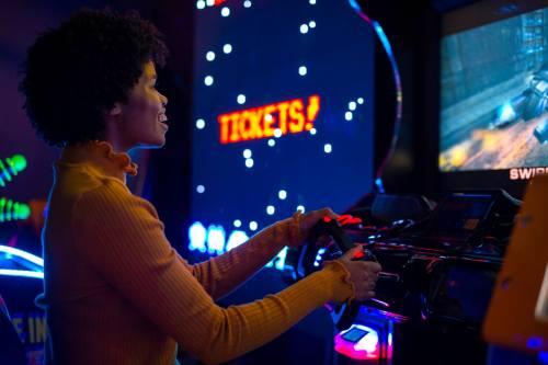 A person with curly hair plays an arcade racing game, focused on the screen with colorful lights in the background.