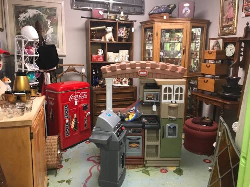 A vintage shop filled with various antiques, including a toy kitchen, retro Coca-Cola fridge, and wooden furniture.