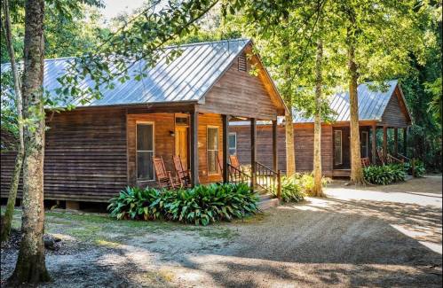 Two rustic wooden cabins with rocking chairs on porches, surrounded by greenery and sunlight filtering through trees.