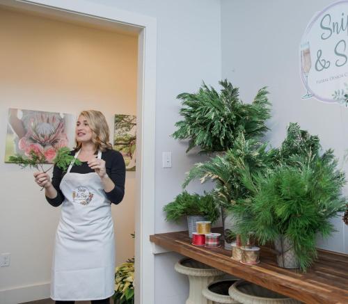 A woman in an apron stands by a table with greenery, demonstrating floral design in a bright, inviting space.