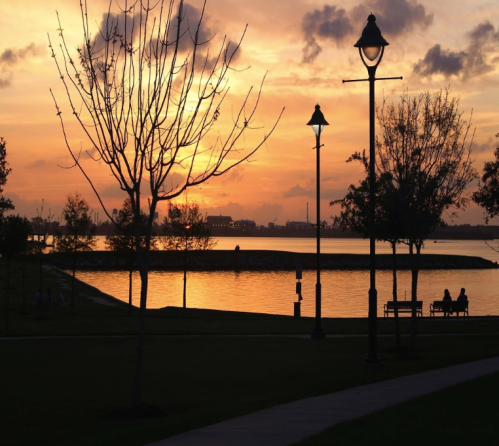 Silhouetted trees and lampposts against a vibrant sunset over a calm lake, with people sitting on a bench.