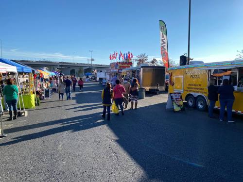A vibrant outdoor market with food trucks and colorful tents under a clear blue sky. People stroll through the area.