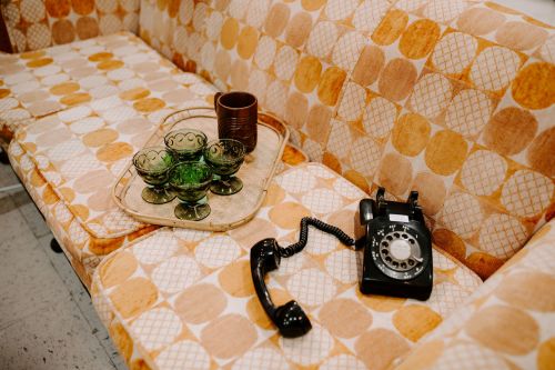 A vintage couch with orange and yellow patterns, a black rotary phone, and green glass bowls on a tray.