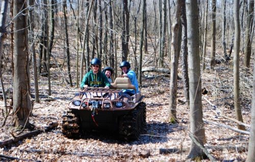 A group of three people riding an ATV through a wooded area with bare trees and fallen leaves.