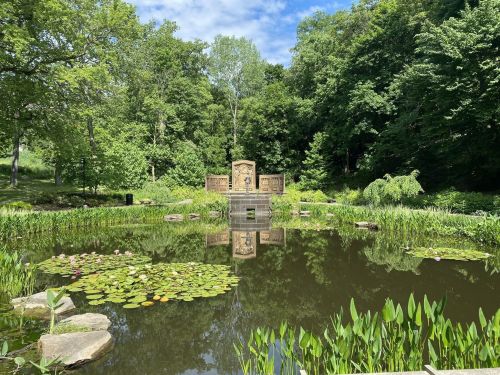 A serene pond surrounded by lush greenery, featuring lily pads and a stone monument reflecting in the water.