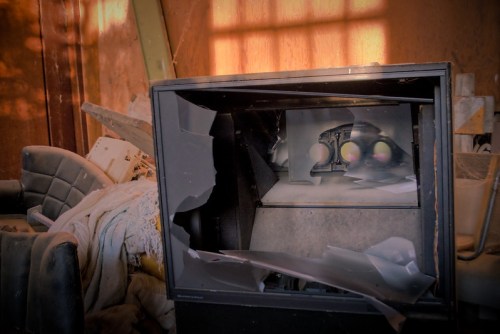 A broken television sits among debris in a dimly lit, abandoned space, with shattered glass and scattered furniture.