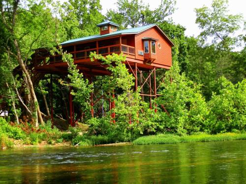 A red treehouse on stilts surrounded by lush greenery and a calm river in the foreground.
