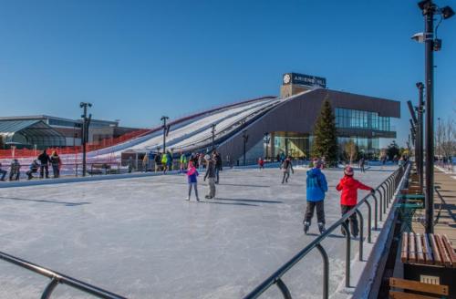 A busy outdoor ice skating rink with skaters, surrounded by a modern building and a snow-covered hill in the background.