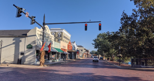 A quiet street scene with shops, traffic lights, and a clear blue sky. A car is driving down the brick-paved road.